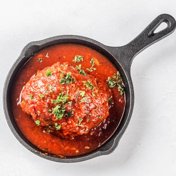 A large Italian meatball covered in rich red marinara sauce and fresh parsley in a cast iron skillet.