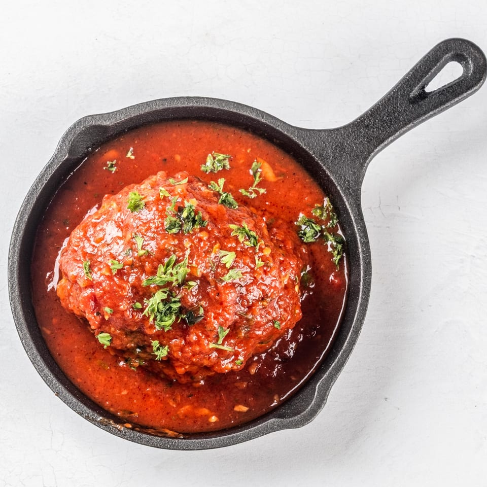 A large Italian meatball covered in rich red marinara sauce and fresh parsley in a cast iron skillet.
