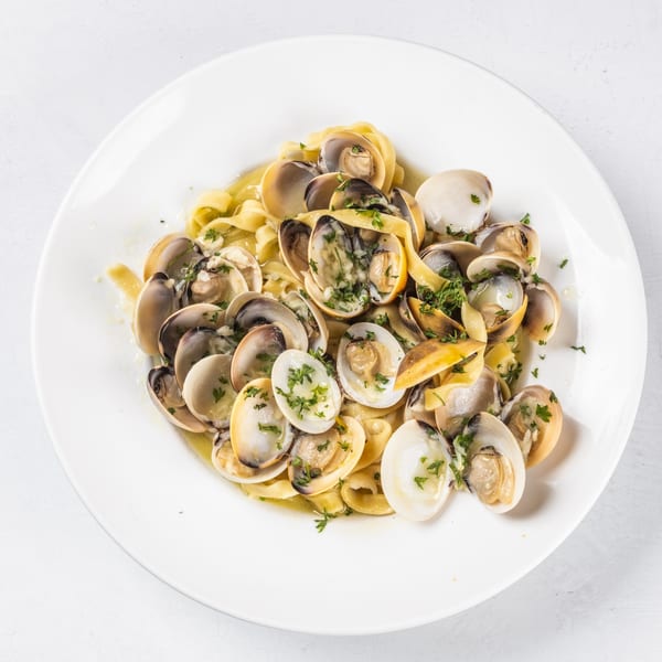 A white plate holds a serving of linguine pasta with clams in a light sauce, garnished with parsley.