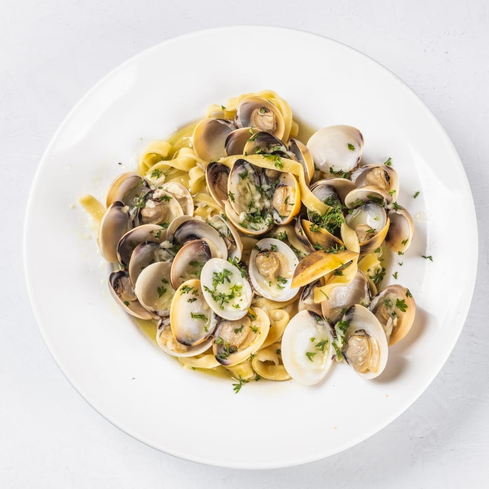 A white plate holds a serving of linguine pasta with clams in a light sauce, garnished with parsley.