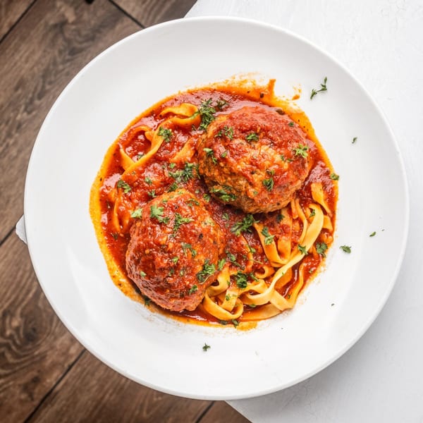 Two large meatballs with red sauce and fettuccine pasta, garnished with parsley on a white plate.