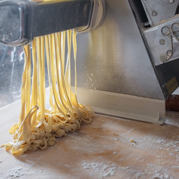Freshly made fettuccine pasta emerges from a pasta machine onto a floured wooden surface.