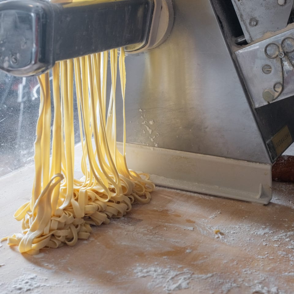 Freshly made fettuccine pasta emerges from a pasta machine onto a floured wooden surface.